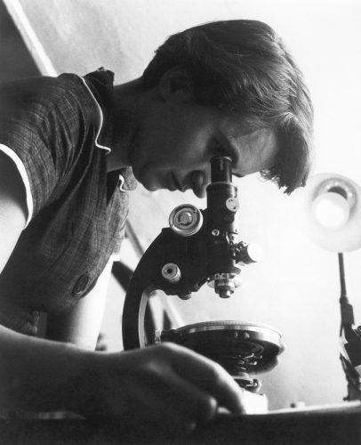 black and white photo featuring a woman with short brown hair, parted at the side, leaning over a microscope